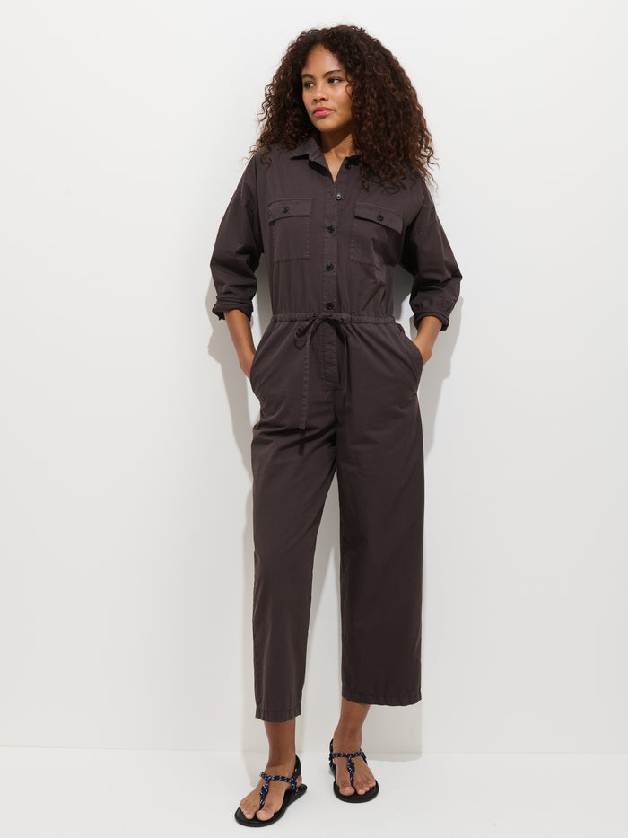 A woman with curly hair wears the Ricard Jumpsuit in Summer Twill—a relaxed fit, dark brown, long-sleeve button-up with front pockets and a waist tie. She stands against a white background, hands in her pockets, looking to the side.