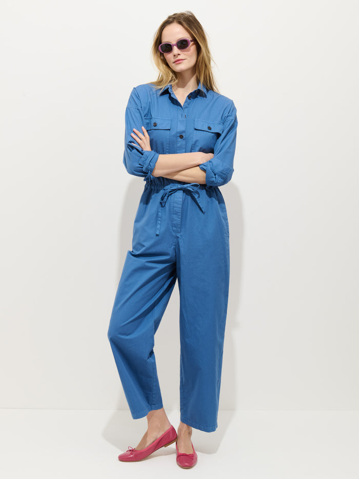 A woman with blonde hair and pink sunglasses stands against a white background, arms crossed, gazing to the side. She wears the Delancey Jumpsuit in Lightweight Twill in blue and pink shoes.