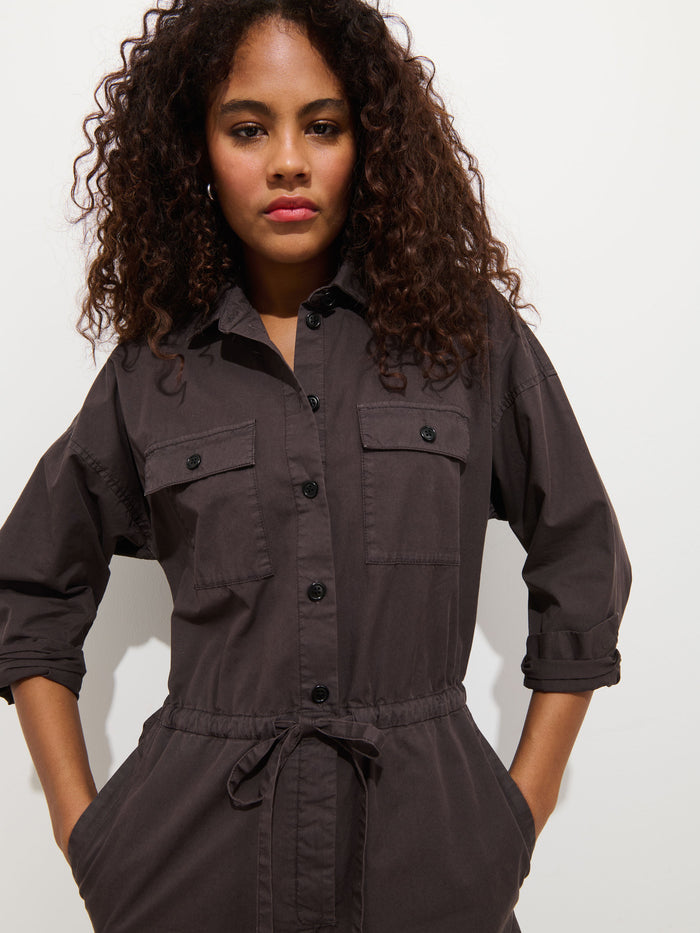 A woman with curly hair stands against a plain white background, confidently wearing the Ricard Jumpsuit In Summer Twill, a relaxed-fit women’s cotton jumpsuit featuring rolled-up sleeves and front pockets.