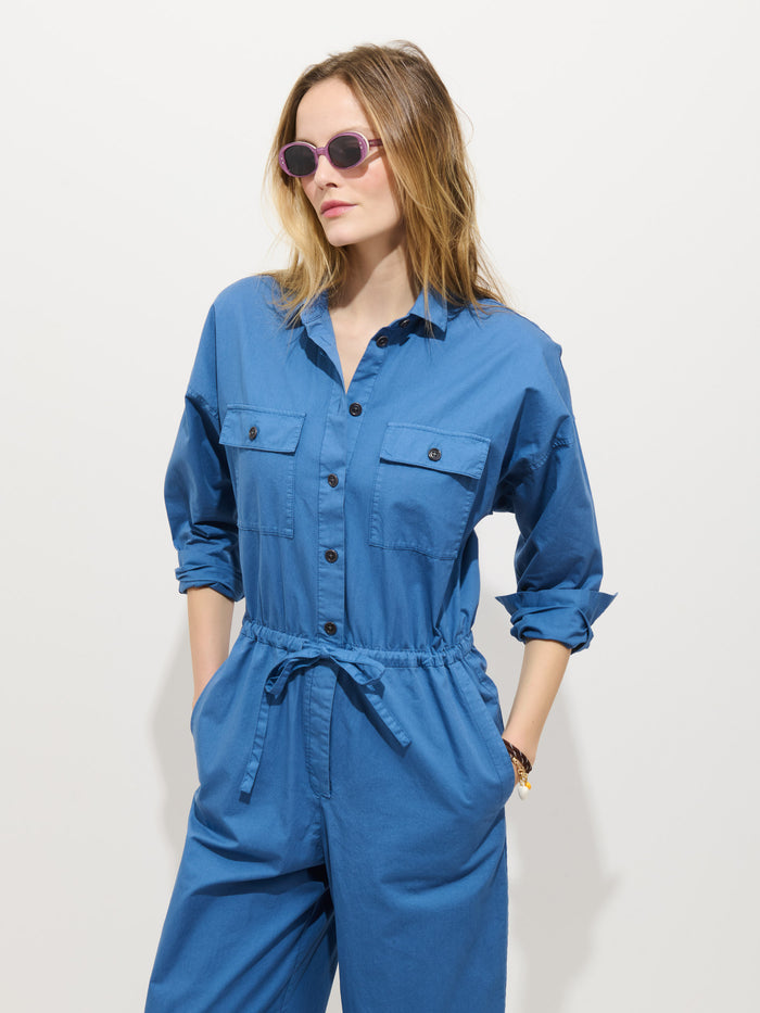A woman in the Delancey Jumpsuit In Lightweight Twill with rolled-up sleeves and purple sunglasses stands against a white background, hands in pockets, gazing slightly to the side.
