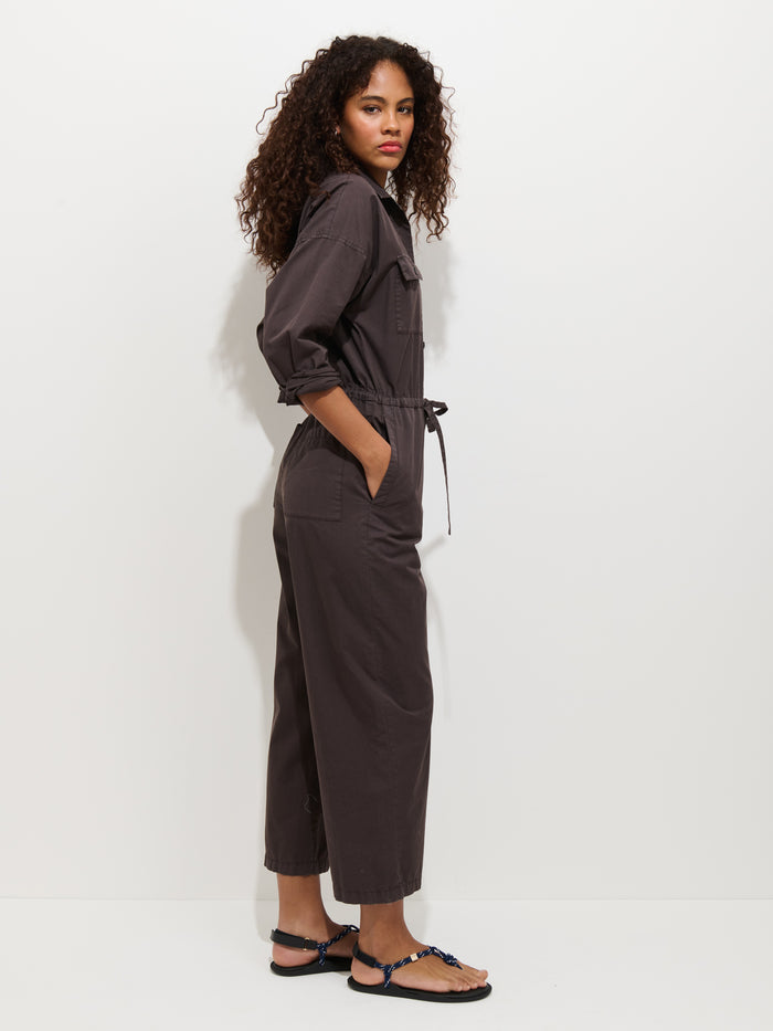 A woman with curly hair stands sideways against a white background, wearing the Ricard Jumpsuit In Summer Twill with rolled-up sleeves and hands in pockets, paired with black sandals.