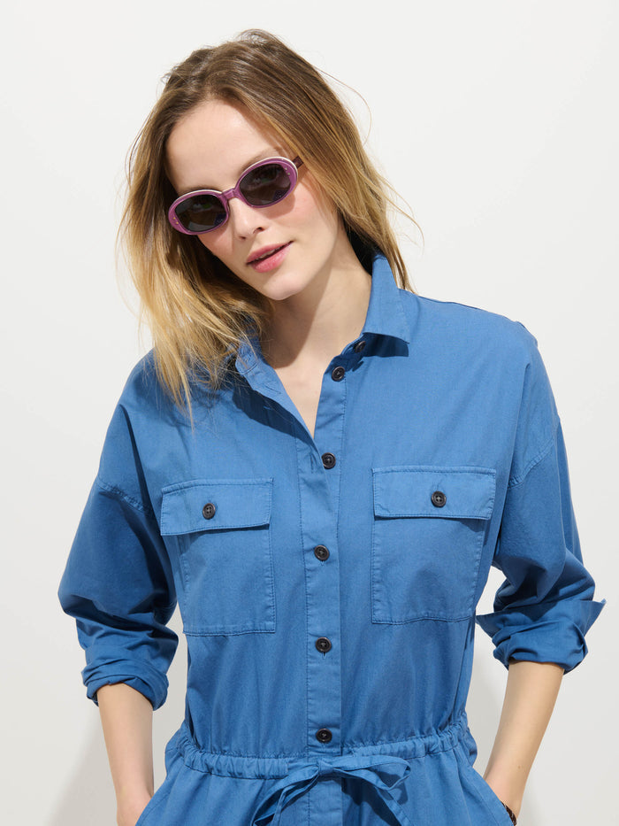 A woman wears the Delancey Jumpsuit in Lightweight Twill with purple sunglasses, standing against a white background with her hands in her pockets and tousled hair.
