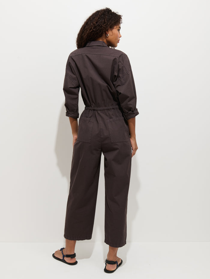 A person with curly hair faces away, wearing the Ricard Jumpsuit in Summer Twill—featuring a relaxed fit, elastic waistband, and wide legs—with black sandals, set against a plain white background.
