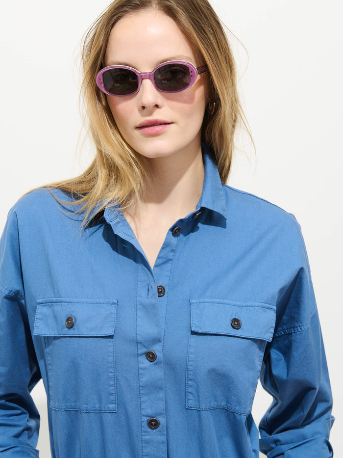 A woman wearing light purple sunglasses and the Delancey Jumpsuit in Lightweight Twill, featuring a relaxed fit and front pockets, stands against a plain white background.