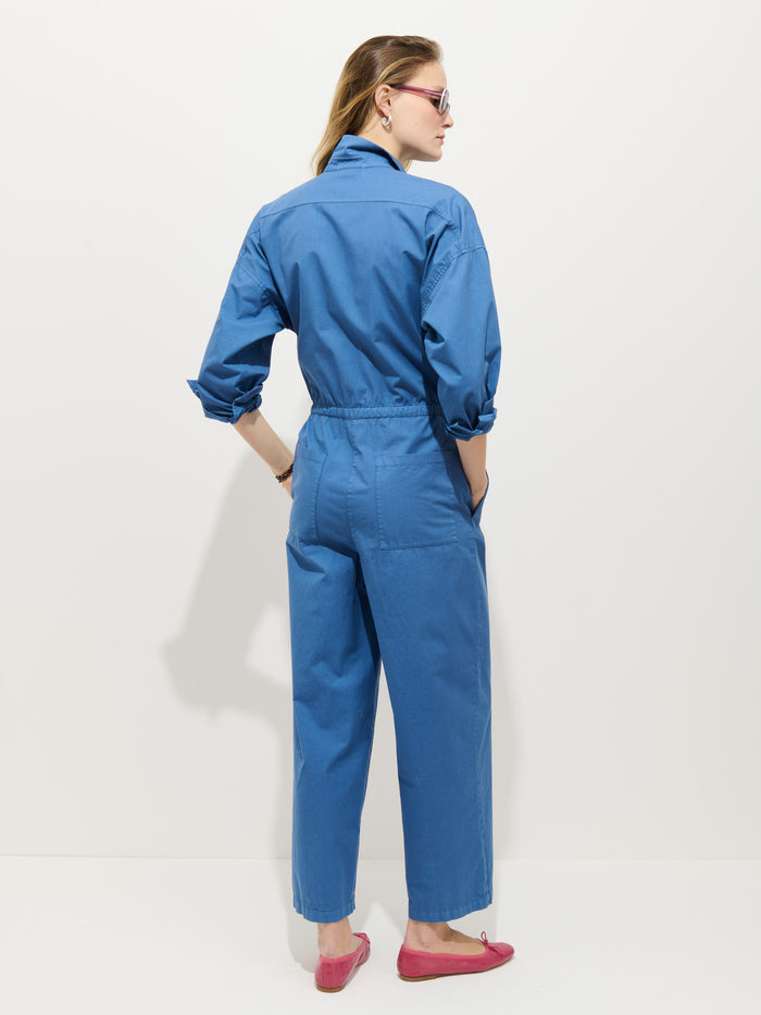 A woman stands facing away in the Delancey Jumpsuit in Lightweight Twill, sleeves rolled up, hands in pockets. She wears pink flats and sunglasses, with light brown hair pulled back, set against a plain white background.