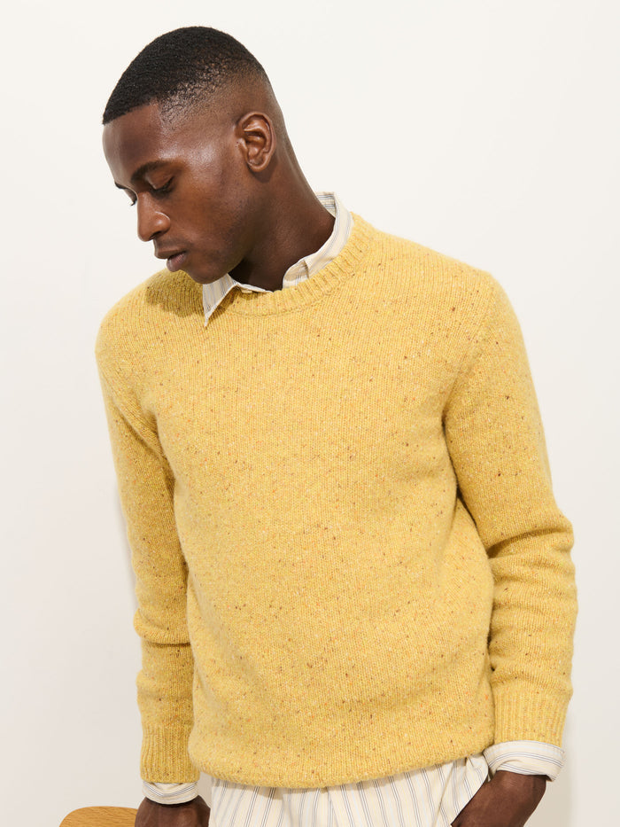 Wearing the Donegal Crewneck Sweater in yellow over a collared, striped shirt, a person stands against a plain light background, looking down with a calm expression.