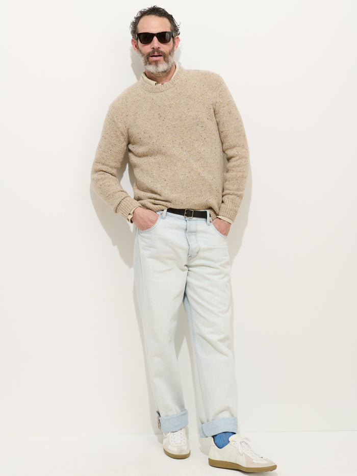 A man with short curly hair, a gray beard, and sunglasses stands against a white wall. He wears a beige Donegal Crewneck Sweater, light blue rolled-up jeans, white sneakers, and has one hand in his pocket.