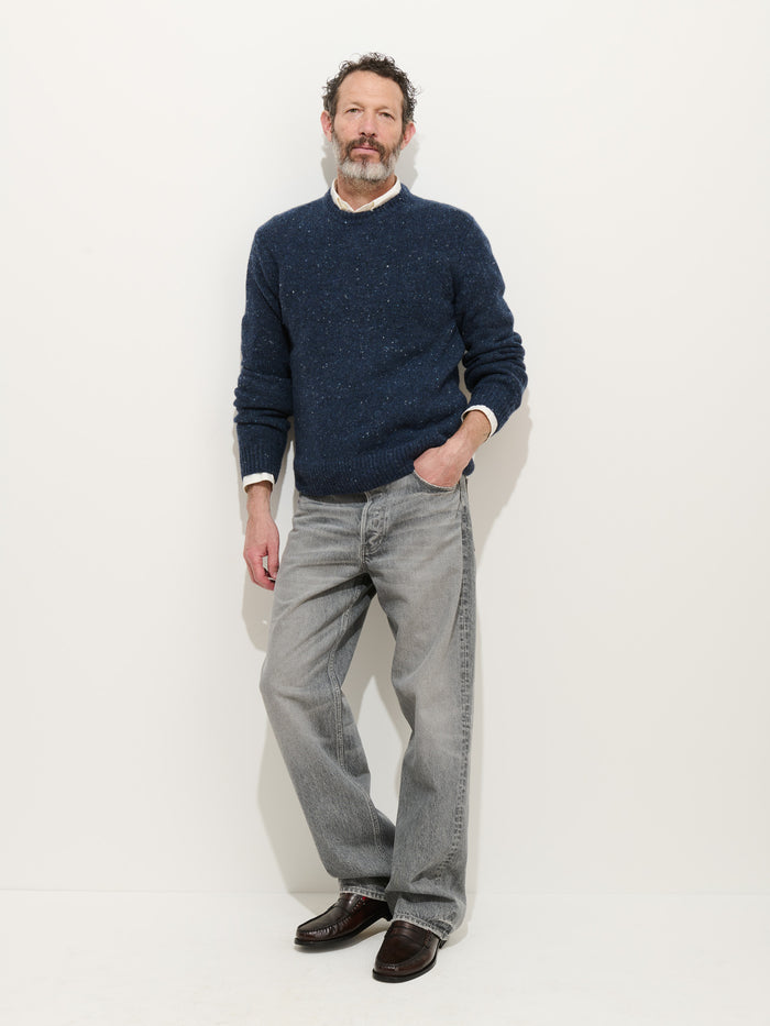 A bearded man with short curly hair stands against a plain white wall, wearing a navy Donegal Crewneck Sweater over a white shirt, paired with light gray jeans and dark brown shoes. One hand is in his pocket as he looks at the camera.