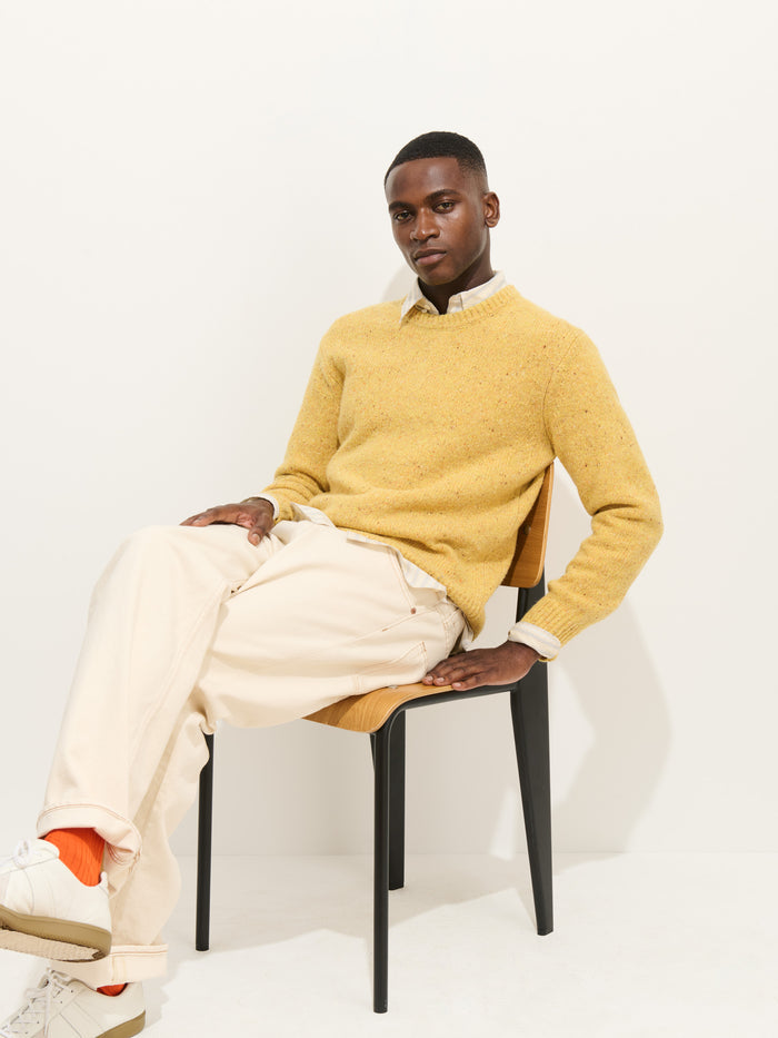 A man sits on a chair against a white background, wearing the Donegal Crewneck Sweater in yellow over a collared shirt, paired with cream pants, white sneakers, and orange socks. He looks at the camera with one leg crossed over the other.