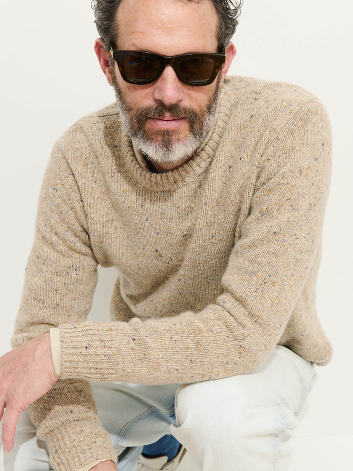 A man with short, curly hair, a beard, and dark sunglasses wears the beige Donegal Crewneck Sweater while crouching and looking at the camera against a plain light background.