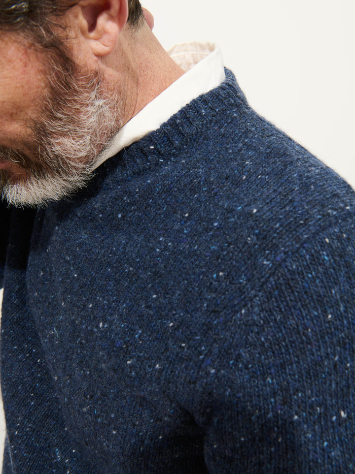 A close-up shows a man in a white-collared shirt layered under a speckled blue Donegal Crewneck Sweater. His salt-and-pepper beard is visible, with his face partly out of frame against a plain, light background.