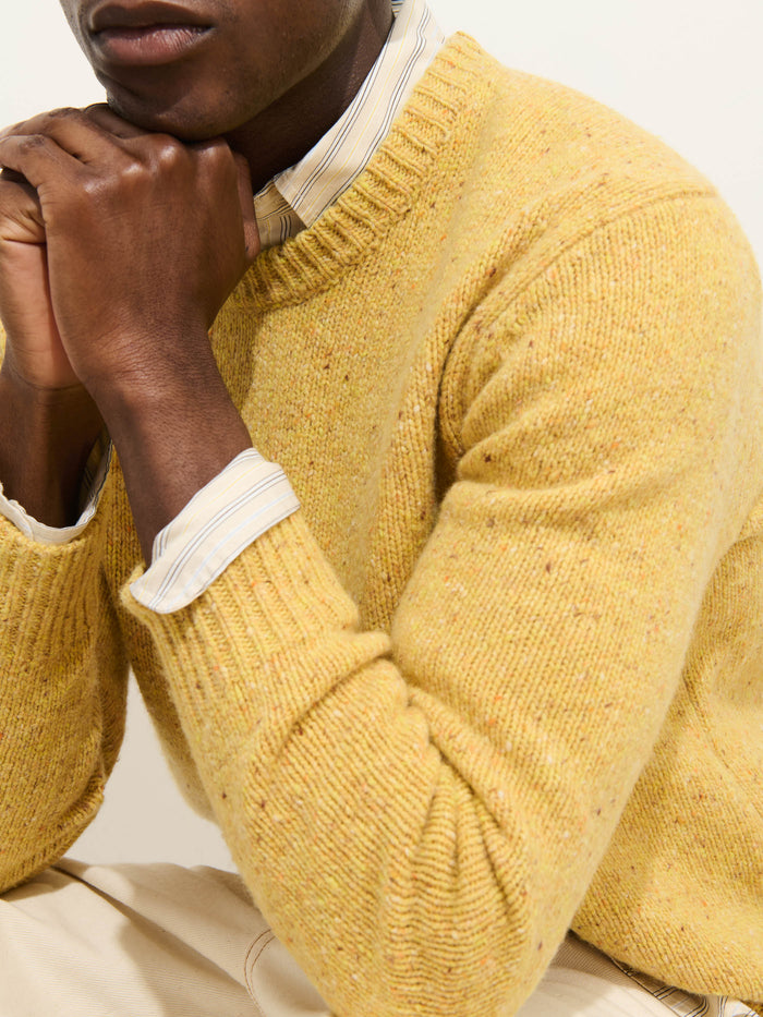 A person with medium brown skin wears the Donegal Crewneck Sweater in yellow speckled knit over a striped collared shirt, hands clasped under their chin. The cropped image shows only the lower face and torso.