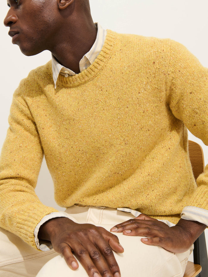 A person sits on a chair, wearing the Donegal Crewneck Sweater in yellow over a light collared shirt and cream pants. Their face is mostly turned away from view.