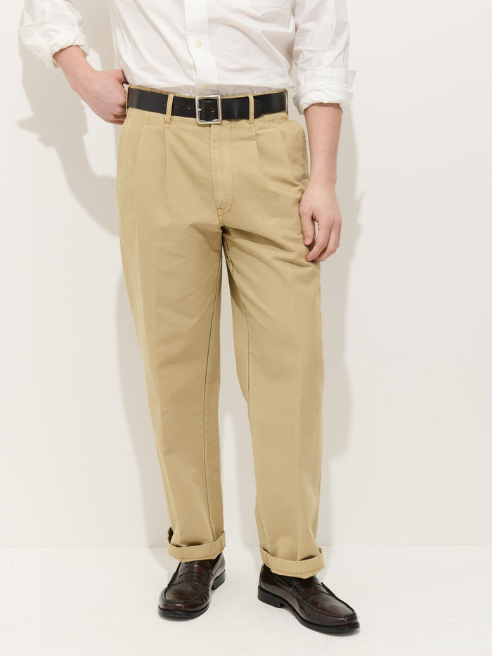 A person wearing a white button-down shirt, Double Pleated Pant In Vintage Twill with a black belt, and dark brown dress shoes stands against a plain light background, left hand in their pocket.