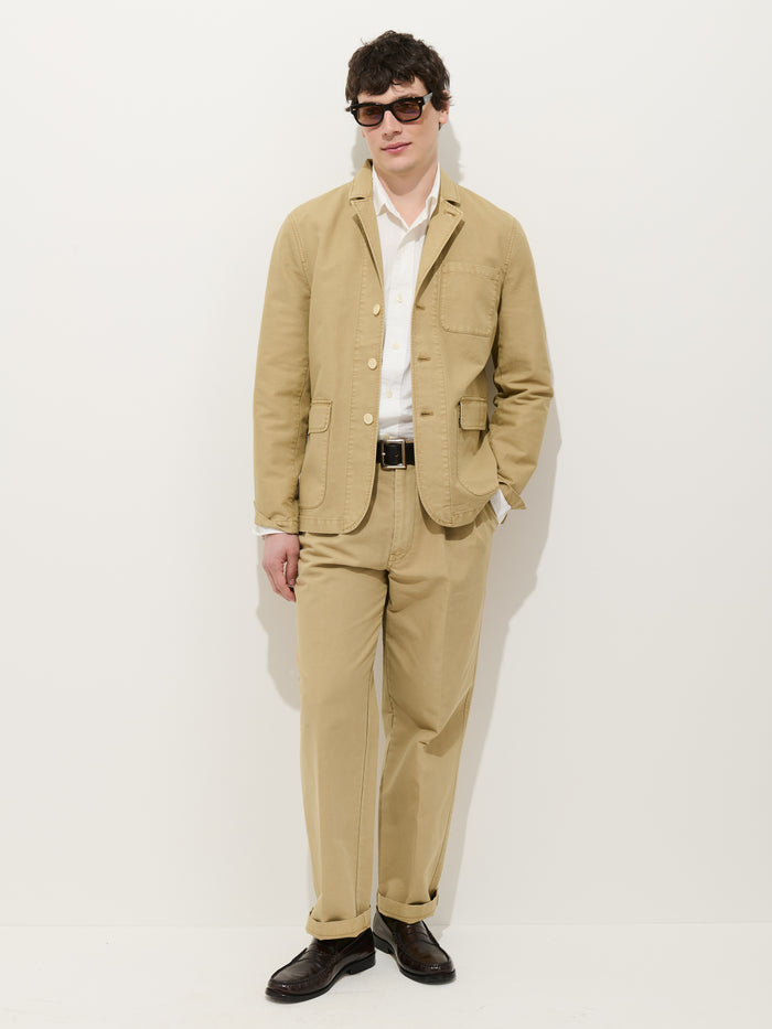 A man with short dark hair, sunglasses, and one hand in his pocket stands against a white background in Alex Mill's Double Pleated Pant In Vintage Twill, paired with a white shirt, dark belt, brown shoes, and tan blazer.