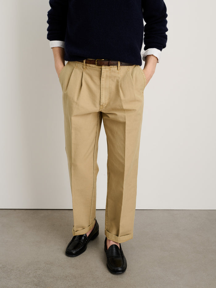 A person stands with hands in pockets, wearing a dark sweater and the Mill Double Pleated Pant in Vintage Twill (tan) with a brown belt and black loafers, against a plain white wall and gray floor. Their face is not visible.