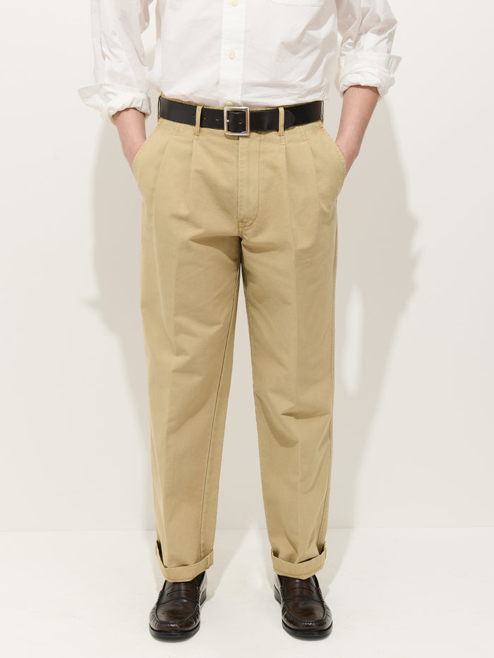 A person stands with hands in their pockets against a plain white background, wearing a white button-down shirt, the Double Pleated Pant In Vintage Twill with straight legs and cuffs, a black belt, and dark brown shoes.