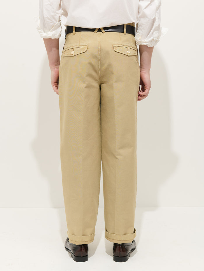 A person faces away, wearing beige Double Pleated Pant In Vintage Twill with buttoned back pockets, a white rolled-sleeve shirt, black belt, and dark brown shoes against a plain white background.