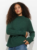 A person with curly hair, clad in the dark green Eastport Rollneck In Cotton sweater and blue jeans, stands against a plain white background, gazing confidently at the camera.