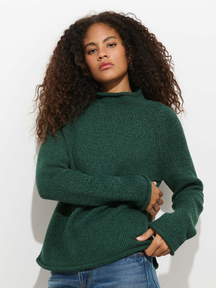 A person with curly hair, clad in the dark green Eastport Rollneck In Cotton sweater and blue jeans, stands against a plain white background, gazing confidently at the camera.