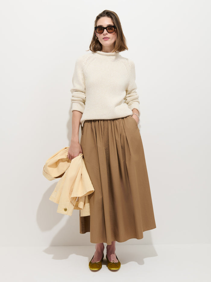 Woman wearing sunglasses, the Eastport Rollneck In Cotton—a cream sweater made from 100% cotton—a long brown skirt, and yellow flats stands against a white background, holding a light yellow jacket in her left hand.