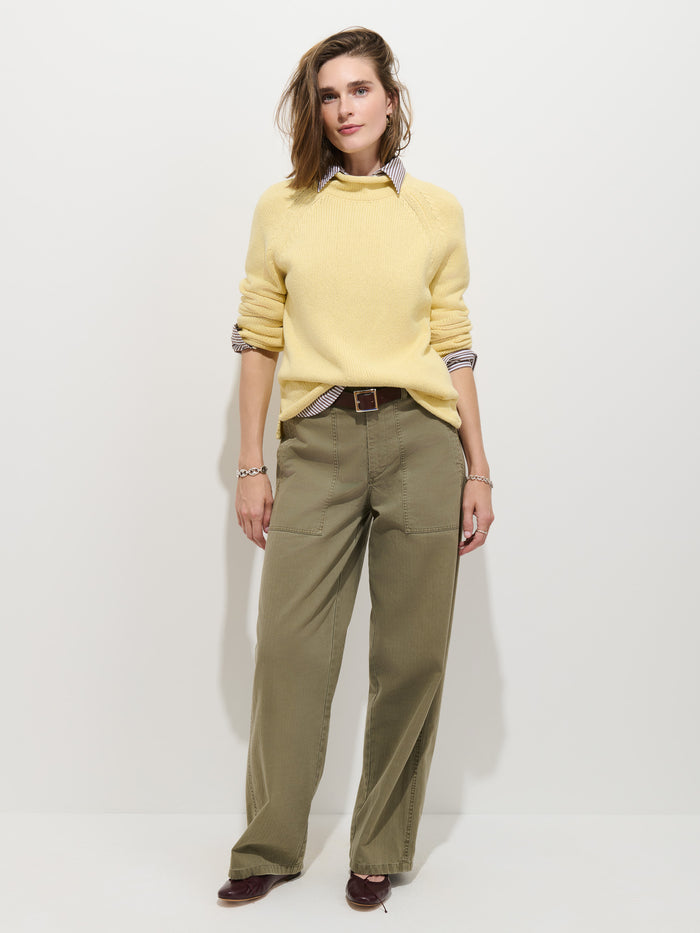 A person stands against a plain white background, wearing the Eastport Rollneck In Cotton—a 100% cotton yellow sweater—over a striped shirt. They pair it with olive green wide-leg pants secured with a brown belt and brown shoes. Their light brown hair frames their relaxed expression.