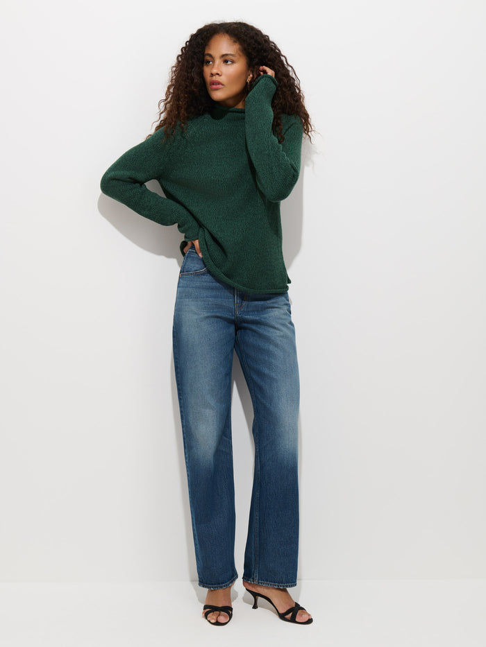 A woman with curly hair stands against a plain white wall, posing with one hand on her hip and the other touching her hair. She is wearing the Eastport Rollneck In Cotton in dark green, blue wide-leg jeans, and black heeled sandals.