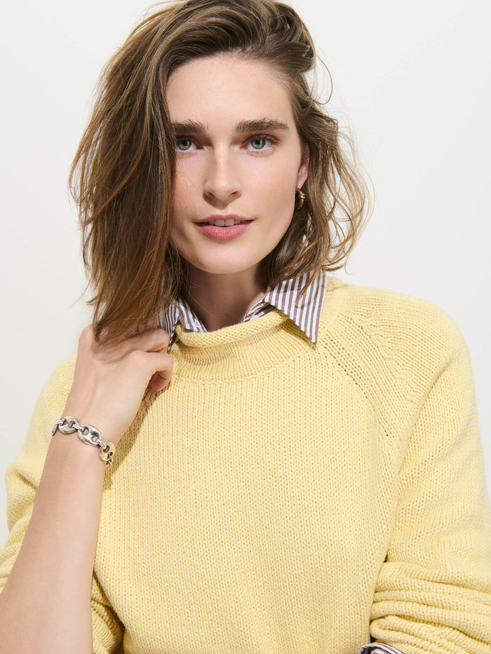 A woman with shoulder-length brown hair wears the Eastport Rollneck In Cotton—a yellow 100% cotton knit sweater—layered over a striped collared shirt, accessorized with a silver bracelet and looking at the camera against a plain background.