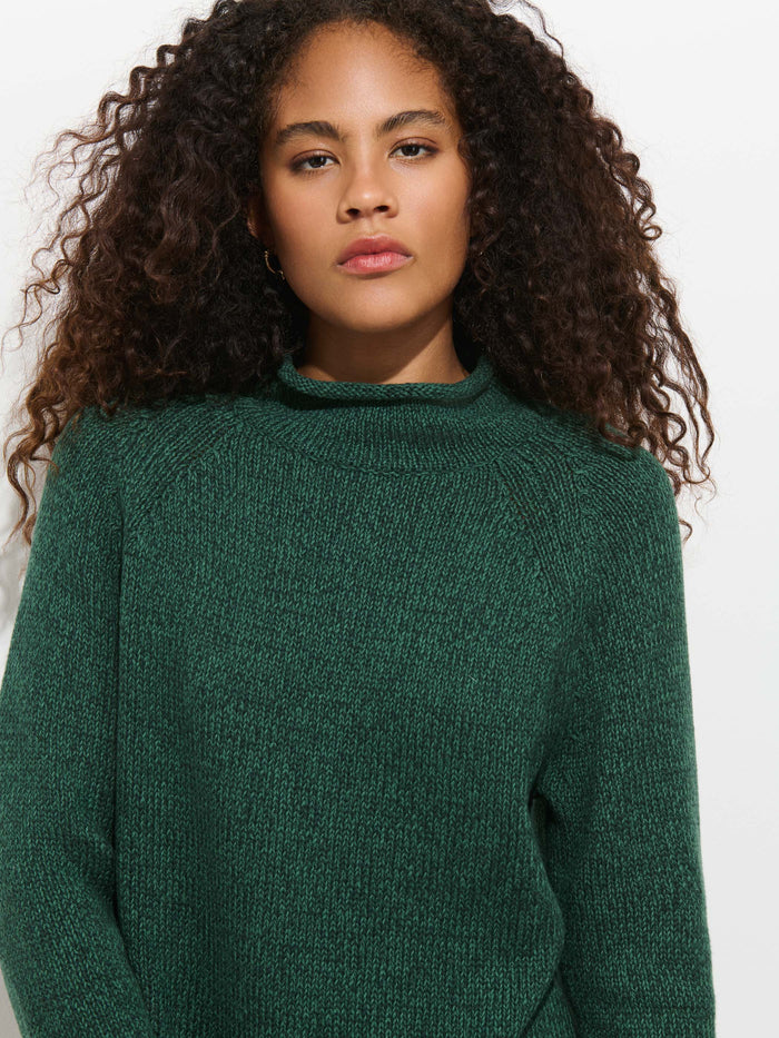 A person with curly brown hair wears the Eastport Rollneck In Cotton, a textured, dark green fisherman’s sweater made from 100% cotton, and looks directly at the camera against a plain light background.