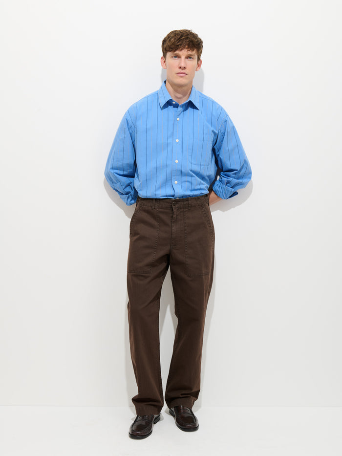 A young man stands against a white background wearing a light blue striped button-up shirt and the Field Pant In Herringbone, vintage-inspired military pants in dark brown, with his hands behind his back and a neutral expression.