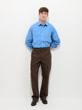 A young man stands against a white background wearing a light blue striped button-up shirt and the Field Pant In Herringbone, vintage-inspired military pants in dark brown, with his hands behind his back and a neutral expression.