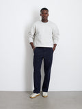 A man stands against a white wall wearing a light gray sweatshirt, Field Pant In Herringbone in dark navy, and white sneakers with light brown soles. He has his hands in his pockets and looks straight ahead.