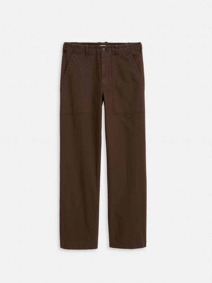 The Field Pant In Herringbone is a pair of garment-dyed brown straight-leg pants, featuring front patch pockets, belt loops, and a button-zip closure, shown flat on a white background.