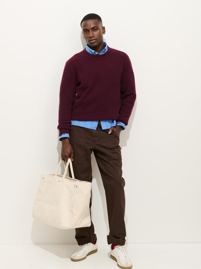 A man stands against a white wall, wearing a burgundy sweater over a blue shirt, the Field Pant In Herringbone in brown, and white sneakers. He holds a large off-white tote bag in his left hand.