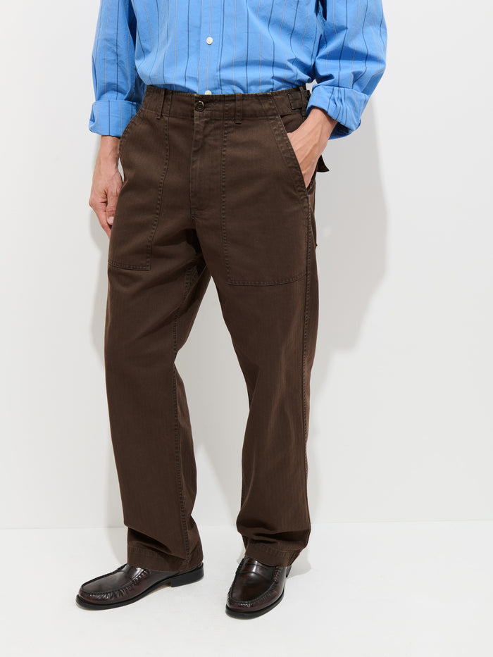A person stands against a white background wearing a light blue button-up shirt, the Field Pant In Herringbone—a vintage-style brown herringbone trouser with large front pockets—and dark brown dress shoes, hands in their pockets.