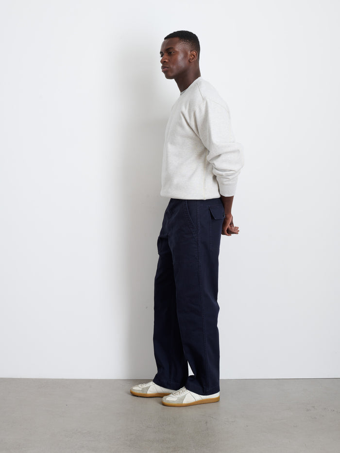 A man stands in profile against a white wall, wearing a light gray sweatshirt and the Field Pant In Herringbone, vintage military style in dark navy, paired with white sneakers. His hands are behind his back as he stands on a gray floor.