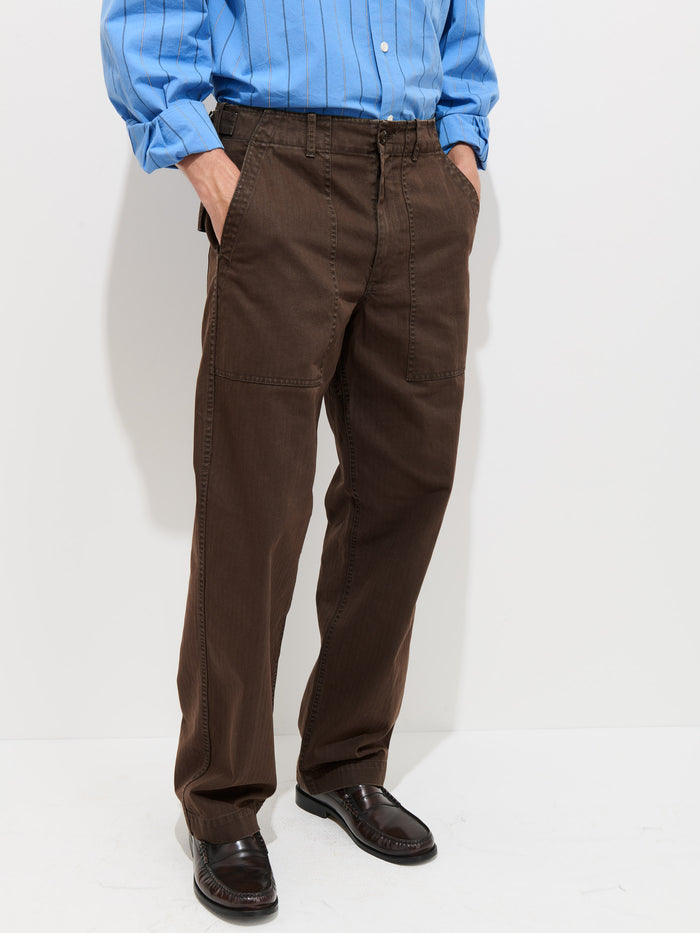 A person stands against a white wall wearing a blue button-up shirt, the Field Pant In Herringbone—a brown, garment-dyed straight-leg style with patch pockets inspired by vintage military pants—and dark brown loafers. Their hands are in their pockets. The photo is cropped at the shoulders.