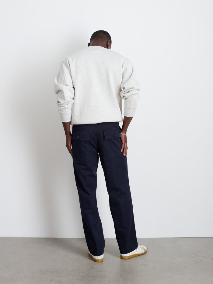 A person faces a white wall with their back to the camera, wearing the Field Pant In Herringbone, a light gray sweatshirt, and white sneakers in a minimal setting with a plain floor and blank background.
