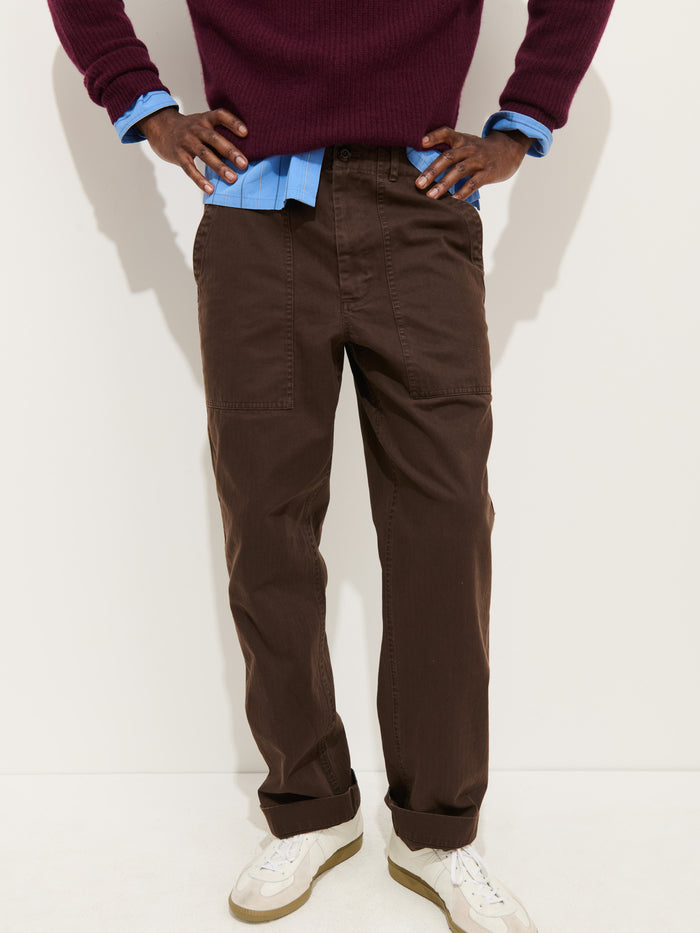 A person wearing the Field Pant In Herringbone in brown, white sneakers, a blue button-up shirt, and a maroon sweater stands with hands on hips against a light background.
