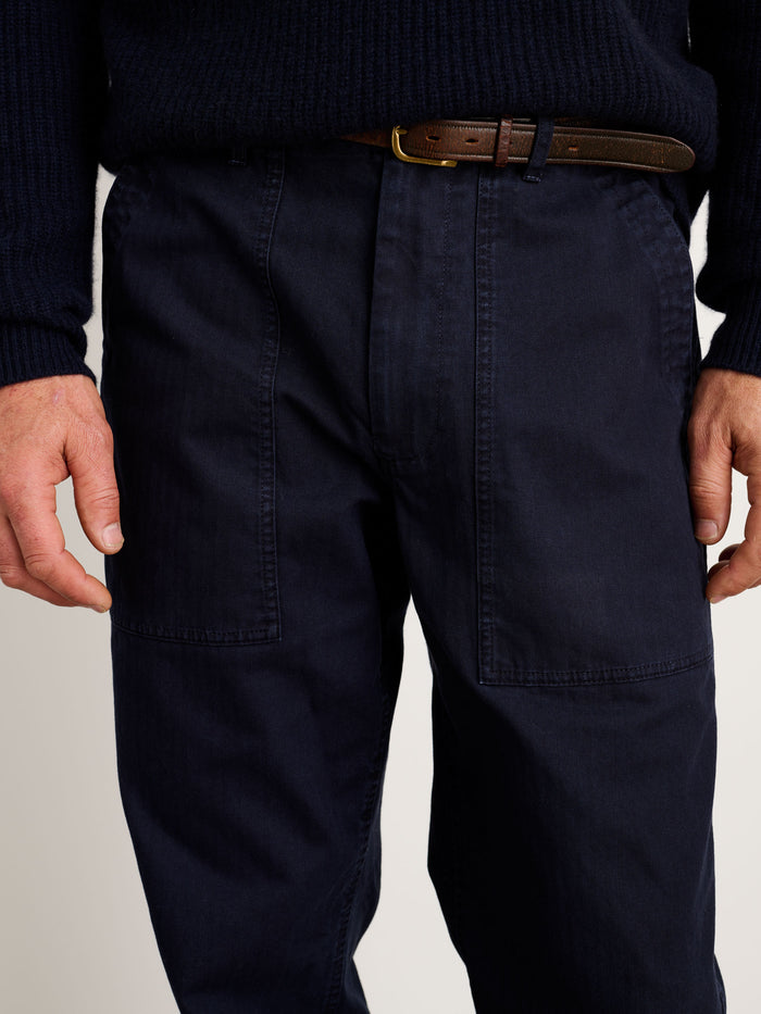 A person wears a dark navy ribbed sweater, Field Pant In Herringbone (navy OG107 style with visible front pockets), and a brown leather belt. The image is cropped waist to mid-thigh, showing relaxed hands at the sides.