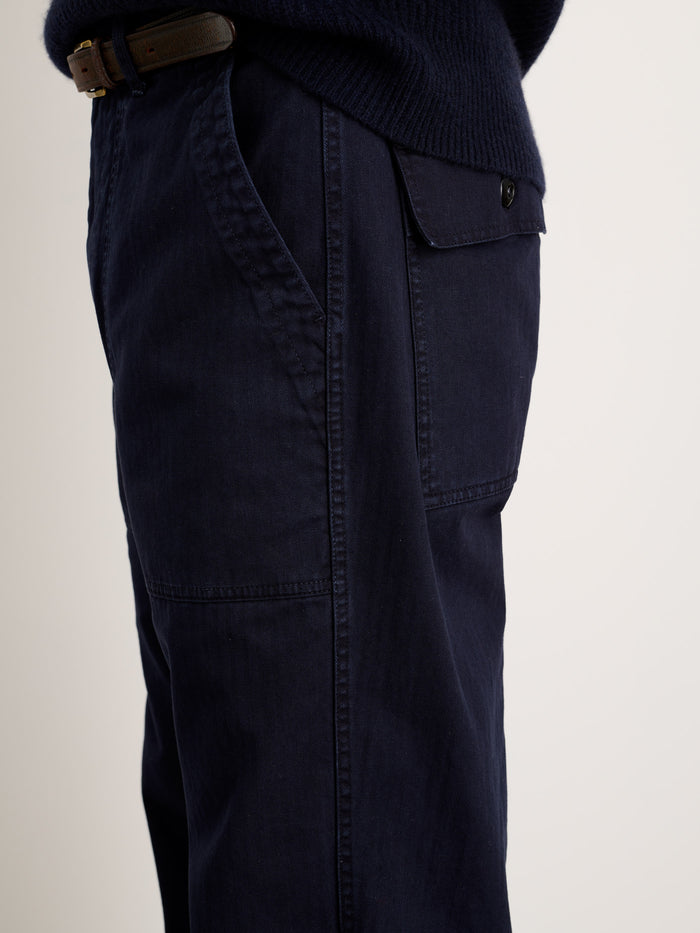A close-up side view of a person wearing the Field Pant In Herringbone, inspired by OG107 military style, in dark navy with a buttoned flap pocket and black belt, paired with a dark sweater against a plain light background.