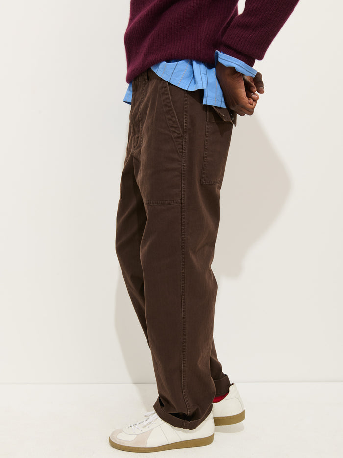A person stands sideways against a plain background wearing the Field Pant In Herringbone, paired with a blue shirt with rolled-up sleeves, a maroon sweater, and white sneakers featuring brown and red accents.