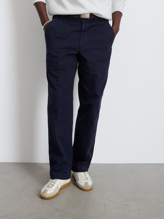 A person stands with hands in pockets, wearing the Field Pant In Herringbone in dark blue, a light gray long-sleeve top, and white sneakers with gum soles. Photo is cropped from shoulders down, against a white wall and gray floor.