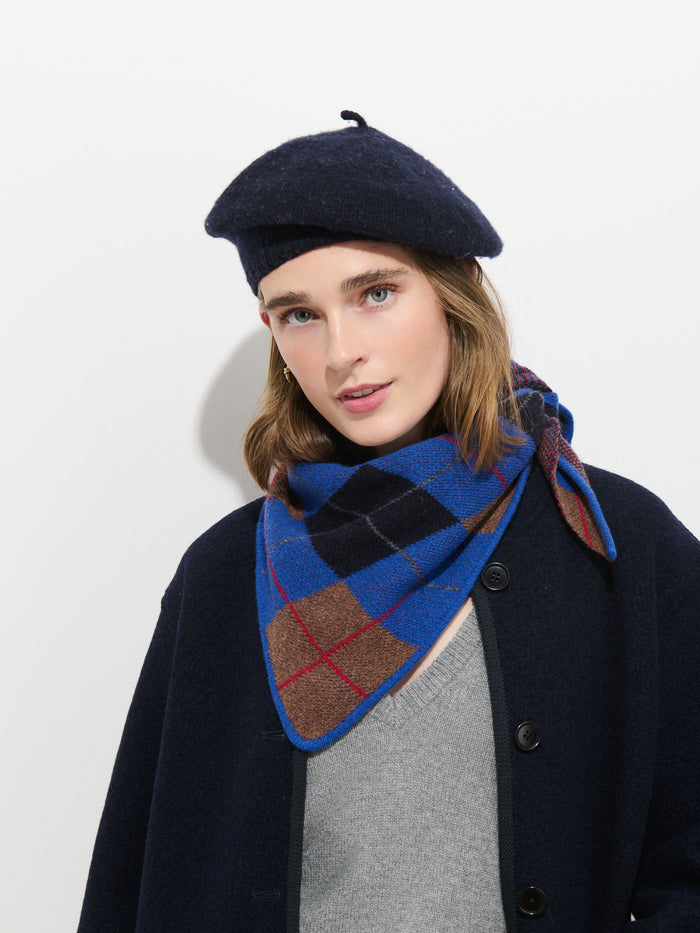 A person wearing a dark beret, a gray top, a dark coat, and the Flynn Argyle Handkerchief Scarf in Wool stands against a white background, looking at the camera.