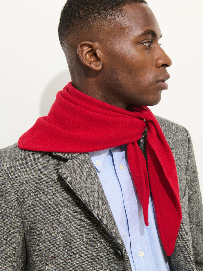 A person wearing a grey coat, light blue shirt, and the Flynn Handkerchief Scarf in Wool in bright red looks to the right against a plain light background.
