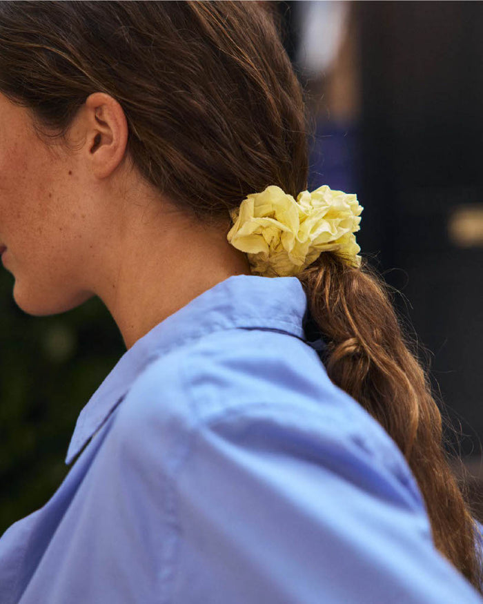 A person with long brown hair tied in a low ponytail with the Frisee Scrunchie In Cotton Poplin, an eco-friendly hair accessory made from recycled cotton, wearing a light blue collared shirt and shown in side profile.