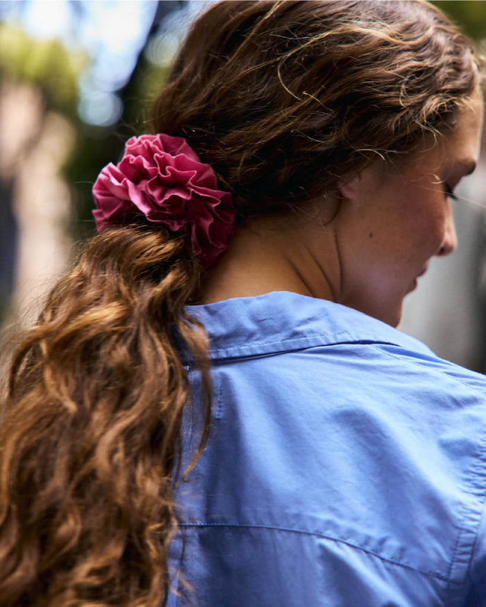 A person with wavy brown hair styled in a ponytail secured by the Frisee Scrunchie In Cotton Poplin, in pink, is shown outdoors from behind and slightly in profile, while wearing a blue shirt.