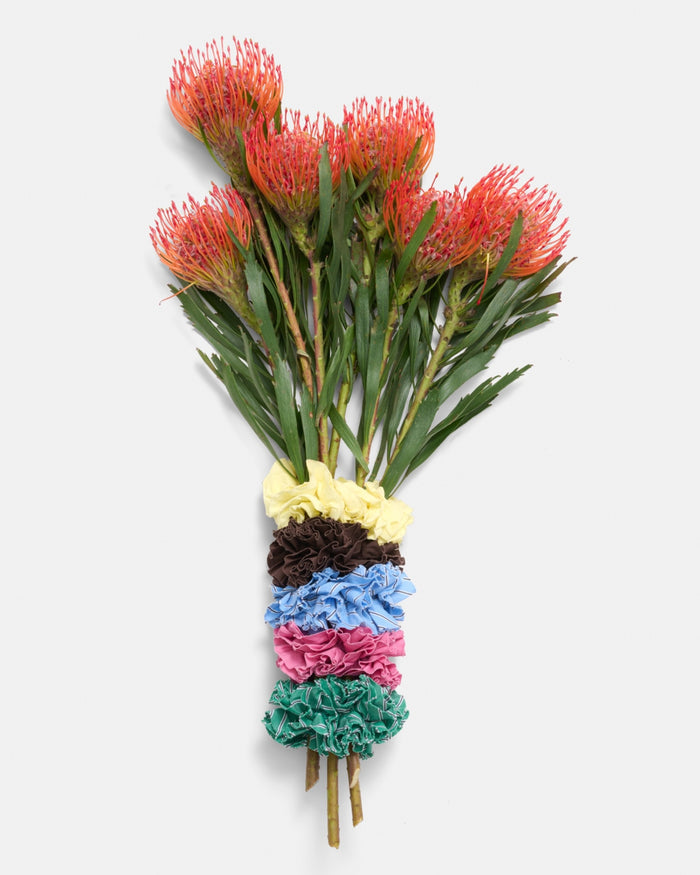A bouquet of orange pincushion protea flowers, with the stems wrapped in six Frisee Scrunchie In Cotton Poplin hair accessories—yellow, brown, blue, pink, turquoise, and green—each crafted from recycled cotton for an eco-friendly touch, set against a plain white background.