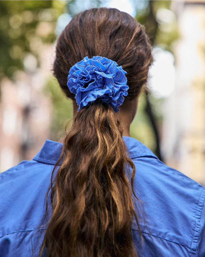 A person with long, wavy brown hair wears the Frisee Striped Scrunchie in Cotton Poplin in blue and a matching blue shirt, standing outdoors with a blurred background of trees and buildings.