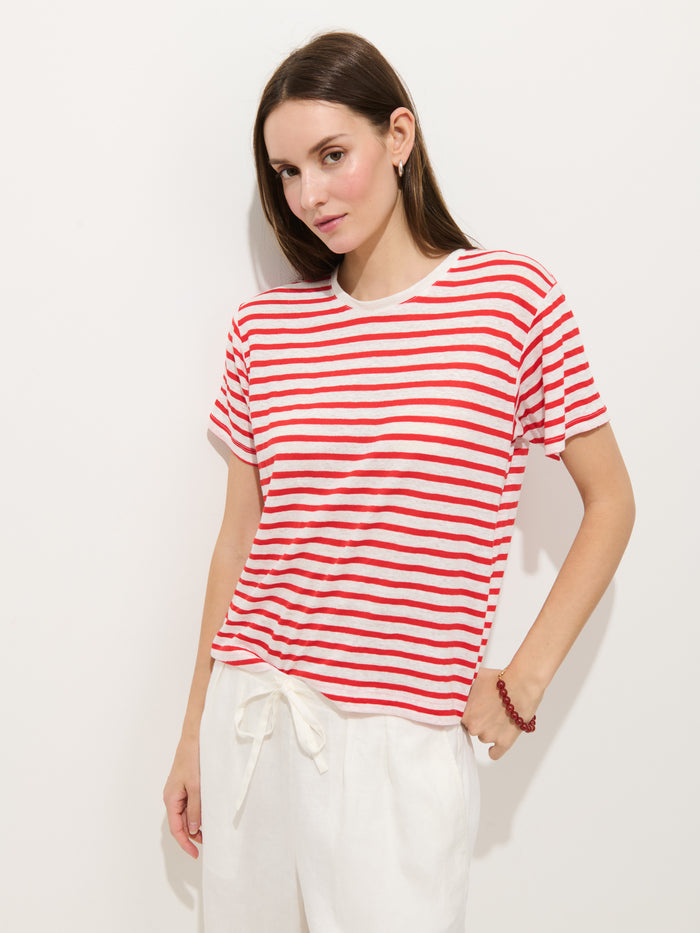 A woman with straight brown hair wears the Garcon Striped Tee In Linen Cotton, featuring red and white stripes, paired with tie-waist white pants. She stands against a plain white background with a neutral expression.